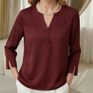 May include: A deep burgundy blouse featuring a V-neckline with vertical pleats, a central placket with four matching buttons, and three-quarter length sleeves with split cuffs and button details, paired with a double-strand white pearl necklace.
