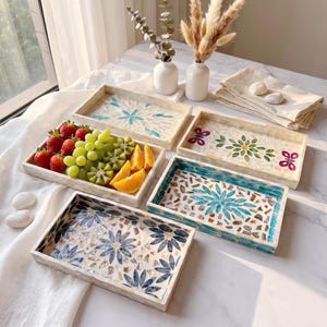 May include: Four rectangular trays with inlaid shell mosaic patterns in blue, green, and pink floral designs. One tray holds fresh strawberries, green grapes, kiwi slices, and orange wedges. Two white ceramic vases with dried stems are nearby.