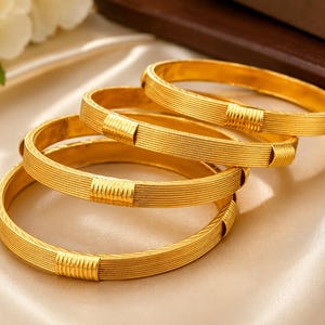 May include: A set of four gold-toned bangles, each featuring a series of fine horizontal lines encircling the band and a single, wider, vertically ribbed rectangular section. The bangles have a polished metallic finish and a circular shape.