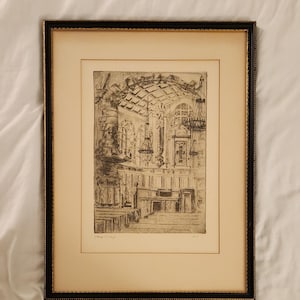 May include: A framed black and white etching depicting the interior of a church with a prominent pulpit, vaulted ceilings, hanging chandeliers, and pews. The artwork is encased in a black frame with a gold inner trim and a cream-colored mat board.