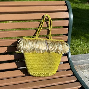 Op de afbeelding: Een chartreusekleurige gehaakte tas met een gestructureerde basis en twee bijpassende handvatten. Het bovenste gedeelte van de tas is versierd met een dikke laag lange, lichtbeige franjes die over de bovenrand van de tas naar beneden hangen.