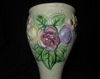 Puede incluir: Un jarrón de cerámica verde pálido con una superficie texturizada y un diseño en relieve de tres rosas en tonos de rosa, amarillo y morado. Las rosas están rodeadas de hojas verdes.
