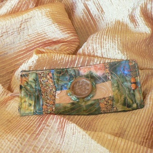 Pode incluir: Uma pequena carteira retangular de tecido patchwork com um botão castanho e um detalhe de contas de madeira. A carteira está sobre um fundo de tecido dourado.