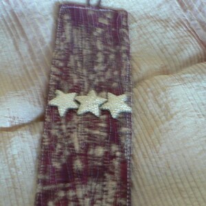 Puede incluir: Un brazalete de tela color burdeos con tres estrellas doradas y un cierre de bot&oacute;n marr&oacute;n.