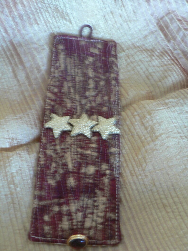 Puede incluir: Un brazalete de tela color burdeos con tres estrellas doradas y un cierre de bot&oacute;n marr&oacute;n.
