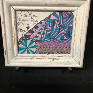 May include: A white framed artwork with a colorful abstract design and the text "Take one day at a time." in black ink.