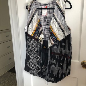 May include: A black and white sleeveless vest with a patchwork design. The vest is made of different fabrics, including a newspaper print, a black and white geometric print, and a black and white cityscape print. The vest has a button closure at the front.