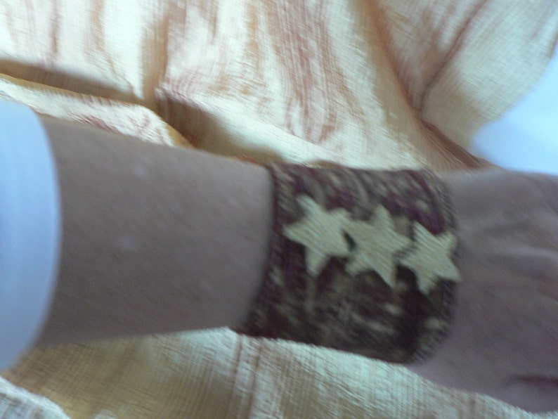 Puede incluir: Primer plano de un brazalete de mu&ntilde;eca de tela marr&oacute;n y dorado con un dise&ntilde;o de estrella. El brazalete se lleva en la mu&ntilde;eca de una persona.