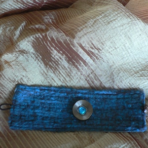 Op de afbeelding: Een tealblauwe stoffen polsband met een zilveren knoop met een turquoise steen.