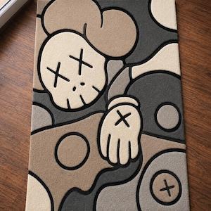 May include: A rectangular area rug featuring a stylized cartoon character design in shades of beige, tan, dark gray, and light gray. The graphic includes a skull-like head with X-shaped eyes, a gloved hand with an X, and circular motifs, all outlined in thick black lines.