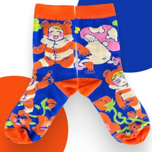 May include: A pair of crew socks featuring a vibrant blue background with an orange cuff. The design displays cartoon figures in orange and white bear-like costumes, pink mushrooms, and orange flowers with green stems scattered across the fabric.