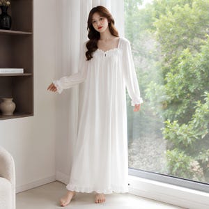 May include: A floor-length white nightgown featuring long sleeves with ruffled cuffs, a square neckline with delicate gathering, and a ruffled hem. The garment is made of a lightweight, opaque fabric and falls in a loose, flowing silhouette.