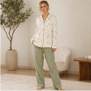 May include: A two-piece pajama set consisting of a long-sleeved, button-down shirt in cream with a delicate green floral print and solid sage green trousers. The shirt features a notched collar, a single chest pocket, and green piping trim.