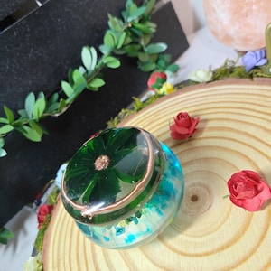 Peut inclure: Une boîte à bijoux ronde en résine avec un couvercle présentant une fleur vert foncé incrustée dans de la résine transparente. La base est bleu translucide avec des éléments décoratifs. Un fil de cuivre entoure le bord du couvercle.