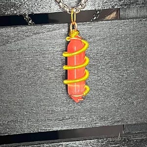 Peut inclure: Un pendentif en pierre orange translucide de forme cylindrique, entouré d'une spirale de fil métallique doré. Le pendentif est suspendu à une chaîne en métal argenté, avec une boucle simple sur le dessus pour la fixation au collier.