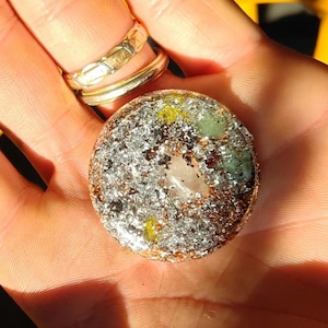 Peut inclure: Un pendentif circulaire en résine contenant un mélange dense de flocons d'argent métallique, de copeaux de cuivre et de petits fragments minéraux incrustés dans des tons vert pâle et jaune, avec un cristal de quartz clair au centre.