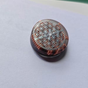 Peut inclure: Un pendentif circulaire en résine brune translucide présentant un motif géométrique argenté en forme de fleur de vie sur la surface supérieure, avec des bobines de fil couleur cuivre visibles incrustées dans la structure en résine.