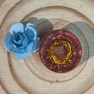 Peut inclure: Un objet en forme de fleur bleu clair avec des pétales superposés, placé à côté d'un bouton rose translucide circulaire présentant un motif texturé complexe et un accent en fil doré enroulé au centre de sa surface.