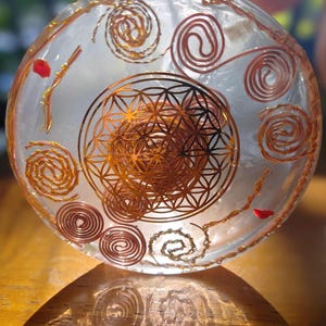 May include: A circular, translucent resin disc containing embedded copper wire coils arranged in spirals. A gold-coloured metal flower of life geometric pattern is centred within the disc, accompanied by small, scattered red fragments.