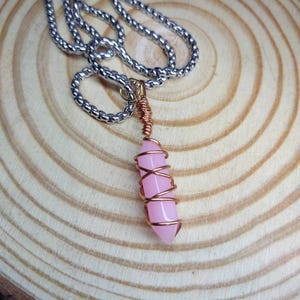 Peut inclure: Un collier composé d'un pendentif en pierre rose clair cylindrique et lisse, entouré de fil de cuivre. Le pendentif est suspendu à une chaîne en métal argenté dotée de maillons arrondis et entrelacés. L'objet est présenté dans son intégralité.
