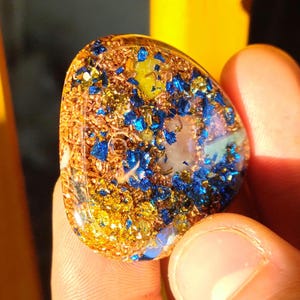 Peut inclure: Un pendentif triangulaire en résine intégrant un mélange de matériaux, notamment des copeaux de cuivre métallique, des éclats de feuille bleue vibrante, des fragments dorés et une pierre translucide centrale sous une finition lisse.