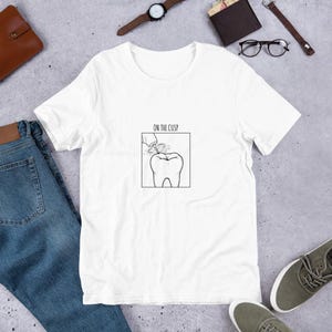 May include: A white short-sleeve t-shirt featuring a black line art graphic of a dental drill working on a tooth inside a square frame. Above the graphic, the text reads ON THE CUSP in a simple, clean, sans-serif font centered on the chest.