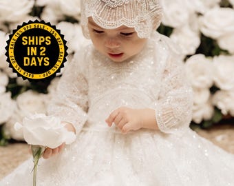 May include: A white long-sleeved dress with intricate lace detailing and a matching lace bonnet. The garment features a full skirt with a subtle shimmer and delicate embroidery throughout the bodice and sleeves, creating a textured appearance.