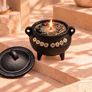 May include: A black three-legged cauldron candle with a lit wick, topped with dried herbs and dark stones. The exterior features a string of gold-colored beads spelling out the text YOUR NAME, accompanied by a matching black lid placed nearby.
