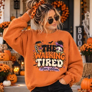 May include: An orange long-sleeve sweatshirt featuring a graphic print with the text THE WALKING TIRED Mom edition. The design includes a skeleton hand holding a pumpkin coffee cup, a cartoon character, bats, and spiderwebs in black and purple.