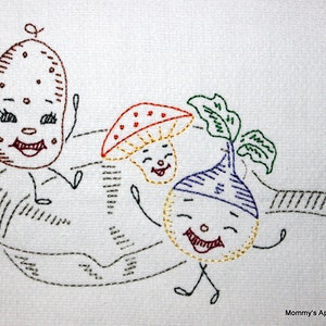 May include: Embroidered white kitchen towel featuring smiling cartoon vegetables: a potato, mushroom, and turnip. The vegetables are dancing in a pan. The towel has the text "Mommy's Apron Strings" in the bottom right corner.