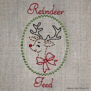 Op de afbeelding: Borduurwerk van een rendier met een rode strik, omringd door een groene cirkel, met de tekst "Reindeer Feed" erboven en eronder.