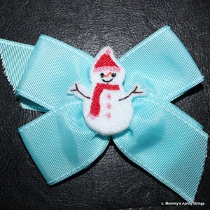 Puede incluir: Un lazo azul claro con una aplicación de muñeco de nieve blanco. El muñeco de nieve lleva un gorro y una bufanda rojos.
