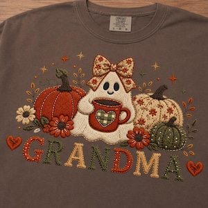 Puede incluir: Una camiseta de manga larga marrón con un diseño bordado de un fantasma con un lazo, sosteniendo una taza roja con un corazón, rodeado de calabazas, flores y estrellas. La palabra GRANDMA está bordada debajo en letras coloridas y estampadas.