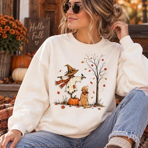 May include: A cream-colored crewneck sweatshirt featuring a graphic print of a scarecrow wearing a sunflower-adorned hat, a golden retriever sitting nearby, a pumpkin, and a bare tree with falling autumn leaves in shades of orange and red.