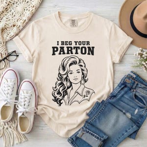 May include: A cream-colored short-sleeve t-shirt featuring a black graphic illustration of a woman with voluminous hair and the text "I BEG YOUR PARTON" printed in a bold, black serif font above the illustration.