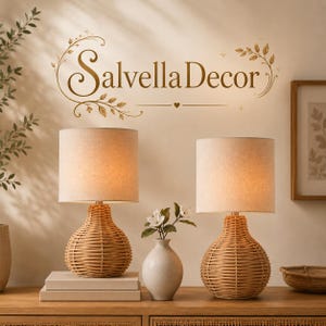May include: Two table lamps with woven light brown rattan bases and cylindrical off-white fabric shades. Between the lamps sits a small white ceramic vase containing a white flower. The text "SalvellaDecor" is displayed on the wall above.