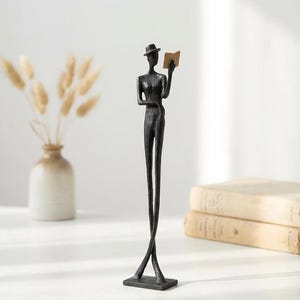 Op de afbeelding: Een hoog, slank, donkerkleurig metalen beeldje van een figuur met een hoed die een open boek vasthoudt. Het figuur staat op een rechthoekige voet met gekruiste benen, heeft een getextureerd oppervlak en houdt een klein, lichtbruin boek in één hand.
