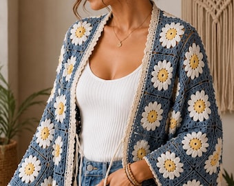 May include: A crochet cardigan featuring a blue square-based pattern with white and yellow daisy motifs. The garment has an open front with a cream-colored border, long wide sleeves, and two hanging cream-colored tassels at the waistline.