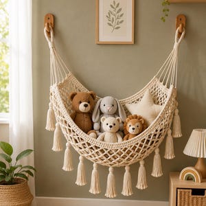 May include: A cream-colored macrame toy hammock featuring a diamond-patterned net design with decorative tassels hanging from the bottom edge. The hammock is suspended by two wooden hooks and contains a collection of plush toys including a bear, a rabbit, a lion, and a star-shaped pillow.