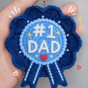 May include: A circular crochet keychain shaped like a rosette award ribbon. The center is light blue with white embroidered text reading #1 DAD, a small red heart, and yellow star accents. The outer border is dark blue with two hanging ribbons.