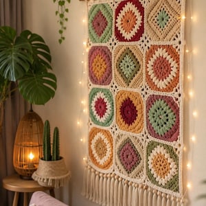 May include: A rectangular crochet wall hanging composed of twelve square motifs in shades of sage green, mustard yellow, terracotta, and cream. The piece is suspended from a wooden rod with a string of small warm white fairy lights and fringe.