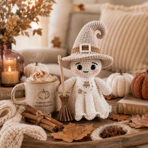 May include: A crocheted ghost doll wearing a beige pointed witch hat with a buckle, holding a small wooden broom. Beside it sits a ceramic mug labeled PUMPKIN SPICE & EVERYTHING NICE, filled with a drink topped with whipped cream and cinnamon.