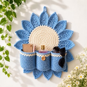 Puede incluir: Un organizador de pared de ganchillo con forma de girasol, con un centro color crema y pétalos azules. Cuenta con tres bolsillos cilíndricos azules con borde de encaje blanco, uno de los cuales está adornado con un botón de madera.