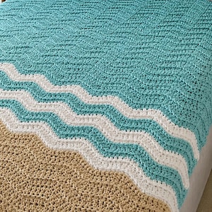 May include: A crochet blanket featuring a wavy pattern in teal, white, and tan. The design mimics a beach scene with a large teal section, a white wave stripe, and a tan base. The texture is chunky and consistent throughout the entire piece.