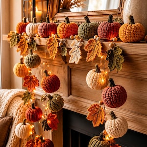 Op de afbeelding: Een decoratieve slinger met gehaakte pompoenen in tinten oranje, rood, groen en crème, afgewisseld met gehaakte herfstbladeren en kleine warmwitte LED-lichtjes, gedrapeerd over een houten schoorsteenmantel met een lantaarn.