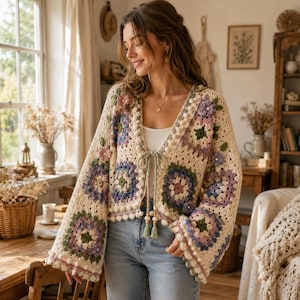 Puede incluir: Un cárdigan de ganchillo color crema con un patrón de cuadrados de la abuela y motivos florales en tonos morado, verde y rosa. La prenda tiene mangas largas y anchas, escote en V y un cierre de lazo frontal con cuentas de madera.