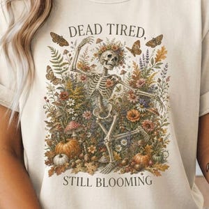 May include: A cream-colored t-shirt featuring a graphic of a skeleton surrounded by colorful autumn flowers, mushrooms, pumpkins, and butterflies. The text "DEAD TIRED, STILL BLOOMING" is printed in a dark font above and below the central illustration.