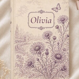 May include: A hardcover book with a cream-colored cover featuring a purple ink illustration of a landscape with wildflowers and a butterfly. The name Olivia is printed in a decorative purple frame at the top center of the book cover.