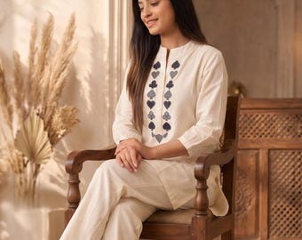 May include: A cream-coloured long-sleeved tunic and matching trousers set. The tunic features a vertical row of dark blue spade-shaped embroidery along the centre front placket. The outfit is paired with simple brown flat shoes.