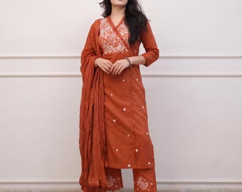May include: A rust-coloured three-piece outfit consisting of a long-sleeved kurta with a V-neckline, matching straight-leg trousers, and a long, draped dupatta. The kurta and trousers feature white floral embroidery accents.